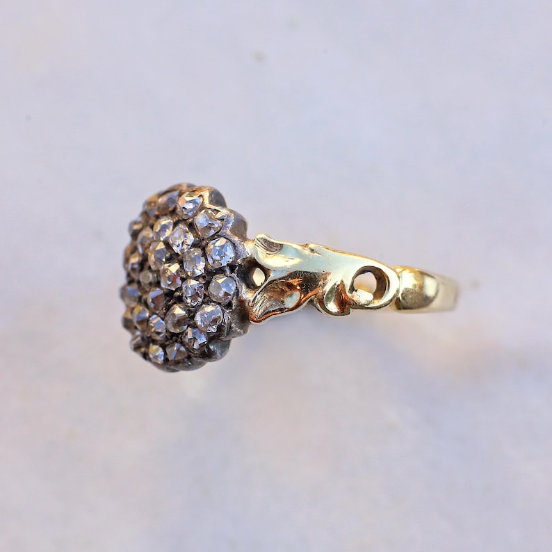 May include: A gold ring featuring a cluster of small, sparkling gemstones. The ring's design includes a decorative floral element and a smooth, polished band. The gemstones are set in a dark metal, contrasting with the gold.