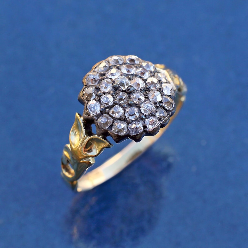 May include: A gold ring with a cluster of small, clear gemstones forming a round shape. The ring has a decorative leaf design on the sides and a gold band. The ring is set against a blue background.