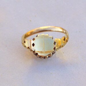 May include: A gold ring with an oval, light-colored center stone. The ring has a decorative, ornate design around the stone and on the band. The ring is set against a light-colored background.