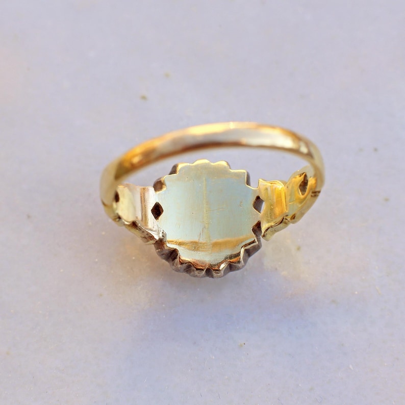 May include: A gold ring with an oval, light-colored center stone. The ring has a decorative, ornate design around the stone and on the band. The ring is set against a light-colored background.