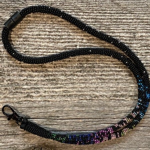 Op de afbeelding: Een zwarte kralenkoord met een regenboogkleurig kralen gedeelte. Het koord heeft een zwarte plastic sluiting en een metalen haak.