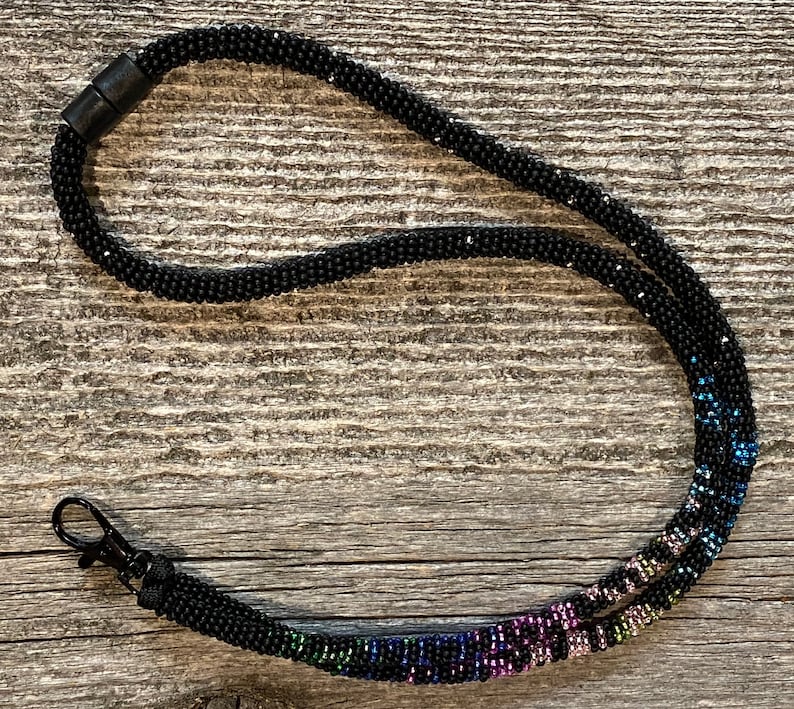 Op de afbeelding: Een zwarte kralenkoord met een regenboogkleurig kralen gedeelte. Het koord heeft een zwarte plastic sluiting en een metalen haak.