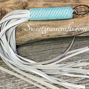 Puede incluir: Un llavero artesanal con un cilindro turquesa con cuentas y un anillo dorado. El llavero presenta una larga borla de tiras de cuero gris claro. El texto "Sweetgrasscrafts.com" es visible.