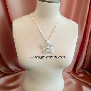 Puede incluir: Un collar con un colgante de estrella de cuentas blancas e iridiscentes verdes y blancas. El collar está en un cordón de satén blanco y se muestra en un busto de maniquí blanco. El texto -Sweetgrasscrafts.com- es visible debajo del colgante.