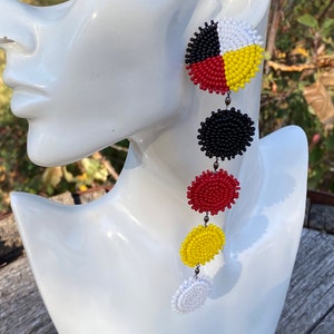 May include: A pair of beaded earrings with a Native American Medicine Wheel design. The earrings are made of black, red, yellow, and white beads. The earrings are hanging from a silver hook.
