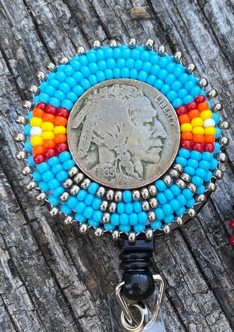 May include: A blue beaded badge holder with a silver-toned buffalo nickel coin in the centre. The coin has the words "Liberty" and "1935" on it. The badge holder has a black retractable cord.