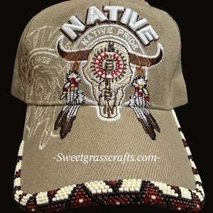 May include: Tan baseball cap with the word "NATIVE" embroidered in white above a bull skull design. The brim is decorated with a beaded pattern in white, brown, and red. The text "Native Pride" is arched above the bull skull.