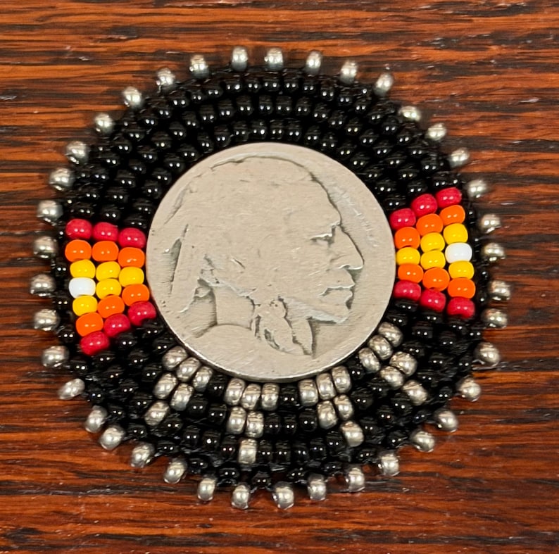 May include: A circular beaded medallion featuring a central silver-toned coin with a profile portrait. The coin is surrounded by concentric rings of small black and metallic silver beads, accented by two triangular patterns of red, orange, yellow, and white beads.