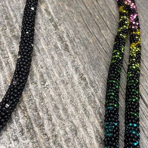 Op de afbeelding: Een zwarte kralenketting met een regenboogpatroon van blauwe, paarse, groene, gele en roze kralen. De ketting is gemaakt van kleine, ronde kralen en heeft een eenvoudig ontwerp.