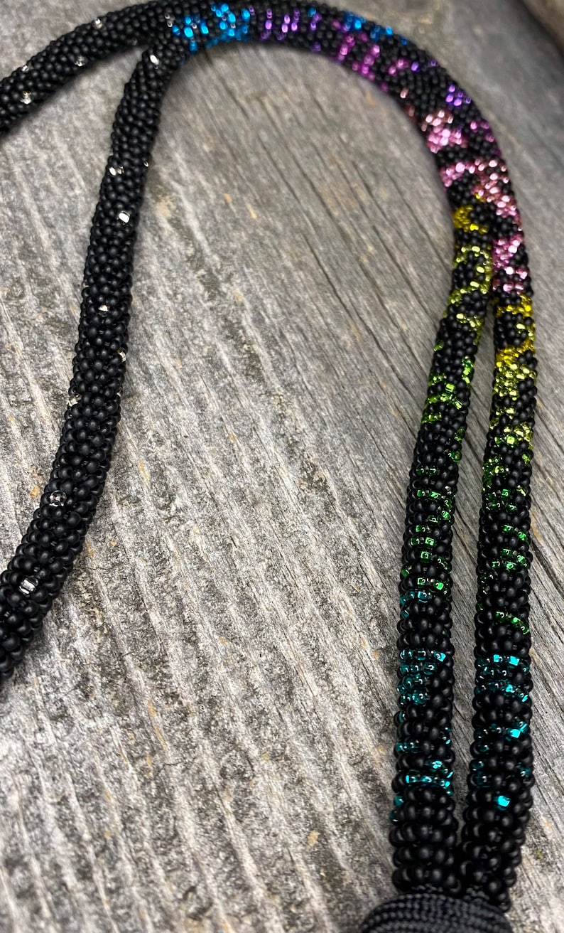 Op de afbeelding: Een zwarte kralenketting met een regenboogpatroon van blauwe, paarse, groene, gele en roze kralen. De ketting is gemaakt van kleine, ronde kralen en heeft een eenvoudig ontwerp.