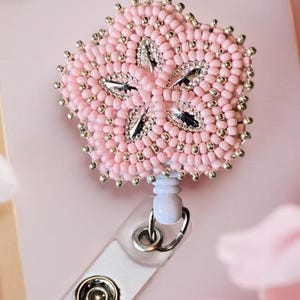 May include: Pink beaded flower badge reel with silver accents and a clear plastic ID holder. The flower design is made of small pink beads with silver beads around the edges. The text "-Sweetgrasscrafts.com-" is visible.