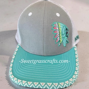 May include: A grey and turquoise baseball cap with a white mesh back. The cap has an embroidered Native American headdress design in turquoise, pink, and yellow. The brim is turquoise with a beaded edge and the text "Sweetgrasscrafts.com".
