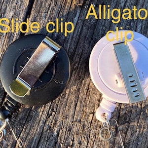 May include: Two circular badge reels, one black and one light pink, each featuring a metal attachment on the back. The black reel has a slide clip, and the light pink reel has an alligator clip. The text "Slide clip" and "Alligator clip" is visible.
