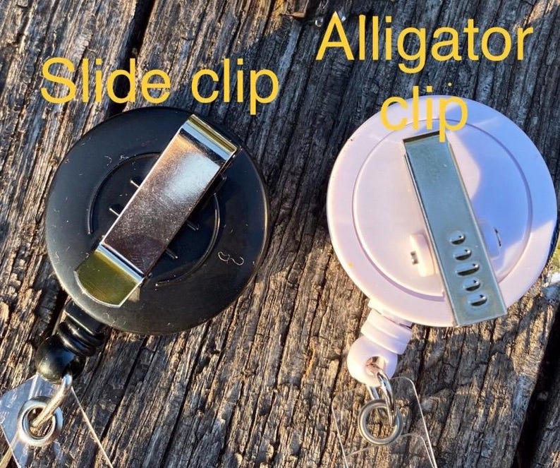 May include: Two circular badge reels, one black and one light pink, each featuring a metal attachment on the back. The black reel has a slide clip, and the light pink reel has an alligator clip. The text "Slide clip" and "Alligator clip" is visible.