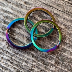 Op de afbeelding: Drie regenboogkleurige sleutelringen in tinten groen, blauw en paars. De ringen zijn van metaal en zijn op elkaar gestapeld.