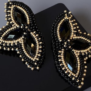May include: A pair of black and gold beaded earrings shaped like butterflies. The earrings are made with small black and gold beads and have a three-dimensional design.