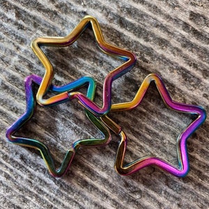 Op de afbeelding: Drie regenboogkleurige stervormige sleutelhangers, elk met een andere kleurverloop, liggen op elkaar gestapeld. De sleutelhangers zijn gemaakt van metaal en hebben een glanzende afwerking.