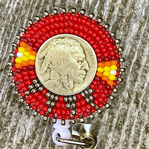 May include: A circular badge reel featuring a 1927 Buffalo nickel coin at the center. The coin is surrounded by concentric rings of red, metallic silver, orange, yellow, and white seed beads. A white plastic clip is attached to the bottom.