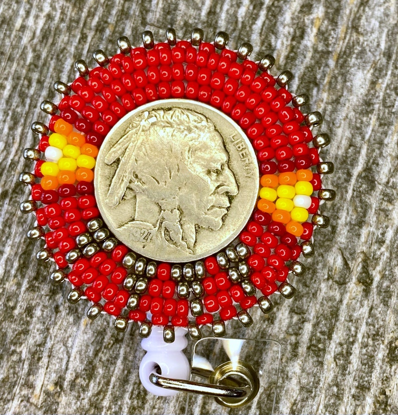May include: A circular badge reel featuring a 1927 Buffalo nickel coin at the center. The coin is surrounded by concentric rings of red, metallic silver, orange, yellow, and white seed beads. A white plastic clip is attached to the bottom.