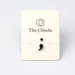 Op de afbeelding: Een paar zwarte puntkomma oorbellen op een witte kaart met de tekst "The Clinda jewelry".