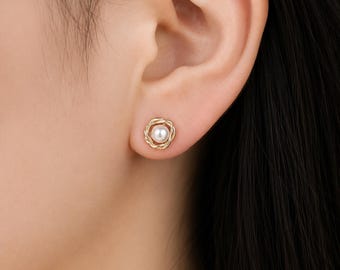 May include: A gold stud earring featuring a central white pearl surrounded by a delicate, twisted metal frame. The circular design creates a textured border around the smooth, spherical pearl, providing a classic and refined appearance for the ear.