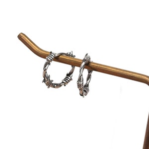 Op de afbeelding: Een paar zilveren creolen met een prikkeldraadontwerp. De oorbellen worden gepresenteerd op een gouden metalen standaard.