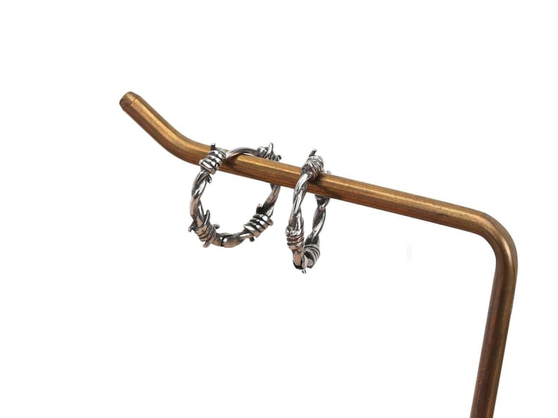 Op de afbeelding: Een paar zilveren creolen met een prikkeldraadontwerp. De oorbellen worden gepresenteerd op een gouden metalen standaard.