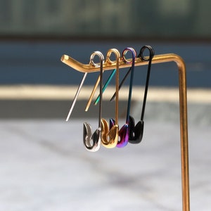 Op de afbeelding: Vijf veiligheidsspeld-oorbellen getoond op een gouden standaard. De oorbellen zijn zilver, goud, teal, paars en zwart.