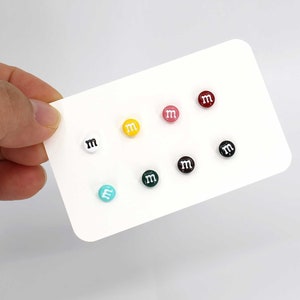 May include: A set of eight colorful stud earrings featuring the letter "M" in white on a variety of colored backgrounds, including red, pink, yellow, green, blue, black, and white.