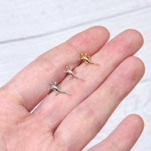 May include: Three small lizard earrings with pink gemstones for eyes. The earrings are shown on a hand, with the gold lizard on the left, the silver lizard in the middle, and the rose gold lizard on the right.