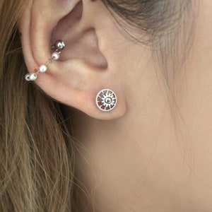 May include: A silver-colored ear cuff and stud earrings. The ear cuff features a small ball at the top, connected by a chain of small white beads. The stud earring is a circular design with a gear-like pattern.