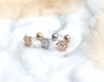 May include: Three surgical steel flower stud earrings with a clear crystal center. The earrings are shown on a white marble surface. The earrings are available in gold, silver, and rose gold.