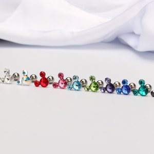 Puede incluir: Una fila de diez pendientes de cristal con forma de Mickey Mouse en varios colores, incluyendo transparente, blanco, rojo, rosa, azul, verde, morado, azul verdoso y negro.