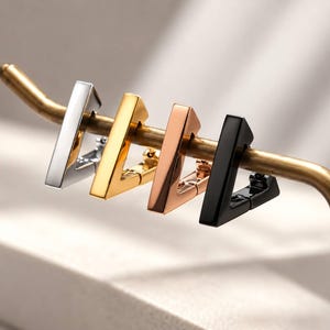 May include: Four triangular hoop earrings in silver, gold, rose gold, and black metal finishes are displayed hanging on a thin, curved, metallic rod. Each earring features a smooth, polished surface and a hinged closure mechanism.