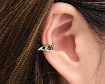 May include: A silver-toned metal ear cuff featuring a series of small, pointed spikes along the outer edge. The accessory is designed to wrap around the cartilage of the ear, providing a secure fit without the need for a piercing.