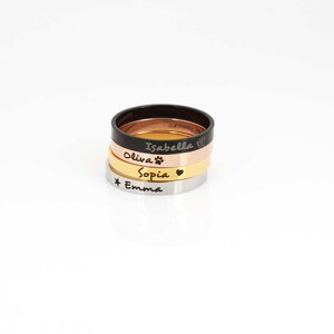 May include: Four stacked rings with different engravings. The top ring is black and engraved with "Isabelle". The second ring is rose gold and engraved with "Olivia" and a paw print. The third ring is gold and engraved with "Sophia" and a heart. The bottom ring is silver and engraved with a star and "Emma".