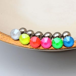 May include: A collection of colorful, faceted beads and shiny silver spheres are arranged along the edge of a cream-colored bowl with a textured, gold-colored rim. The beads are in various bright colors.