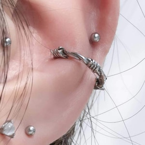 Op de afbeelding: Close-up van een oor met meerdere piercings. Het oor is gepierct met een zilveren oorring met een gedraaid ontwerp, een zilveren oorbel en een zilveren bolvormige oorbel.