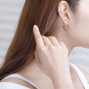 May include: A woman wearing a white tank top with a silver hexagonal hoop earring in her left ear.