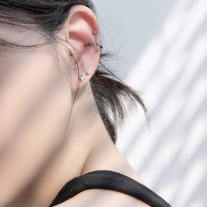 Op de afbeelding: Een close-up van iemands oor met meerdere piercings. Het oor is gepierct met een zilveren oorring, een zilveren oorbel en een zilveren barbell piercing.