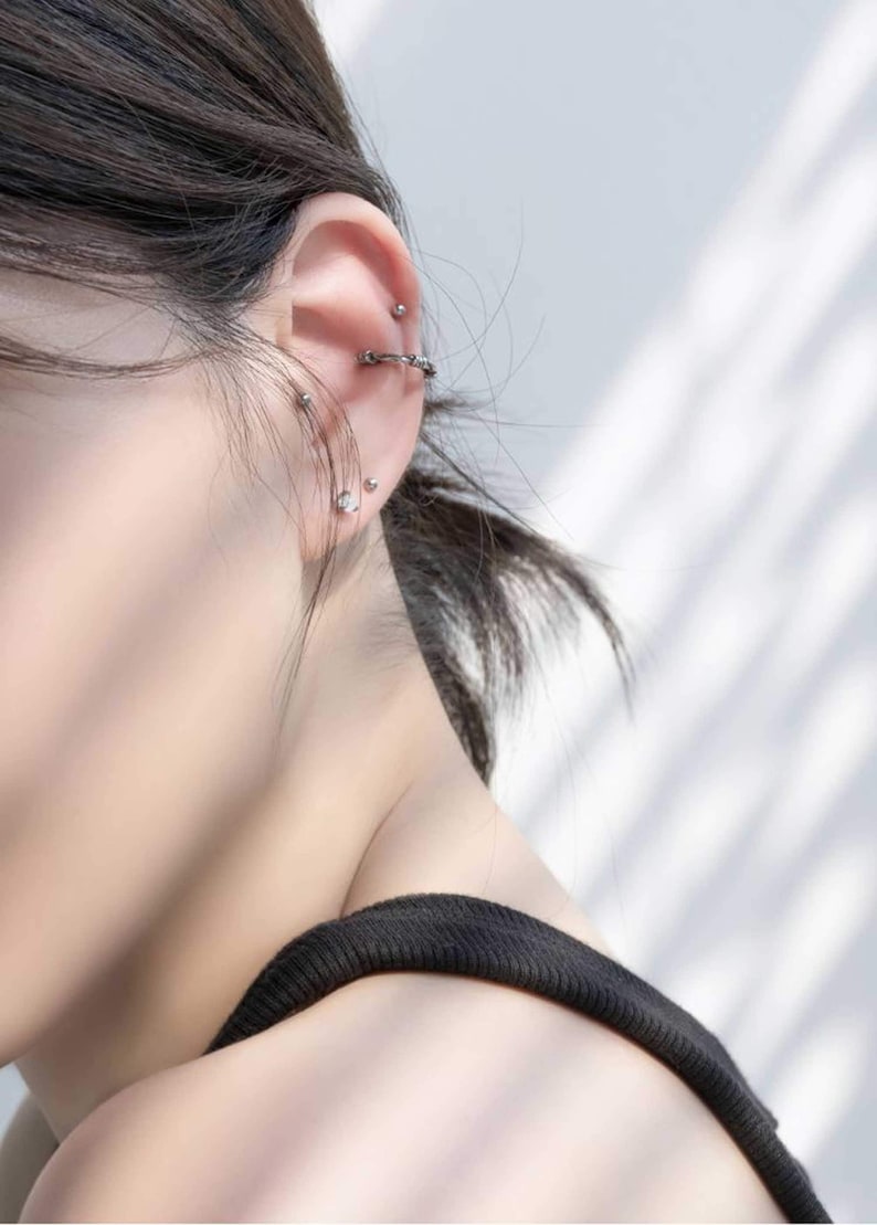 Op de afbeelding: Een close-up van iemands oor met meerdere piercings. Het oor is gepierct met een zilveren oorring, een zilveren oorbel en een zilveren barbell piercing.