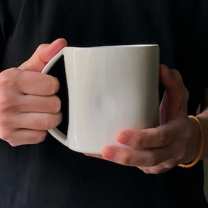 May include: A white ceramic mug with a unique, curved handle. The mug is held in the hands of a person.
