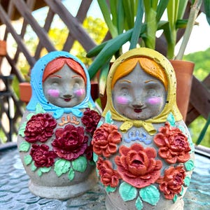 May include: Two decorative Matryoshka dolls. One doll is wearing a blue headscarf, the other a yellow one. Both dolls have red hair, pink cheeks, and are adorned with red rose floral designs. The dolls are made of a gray material.