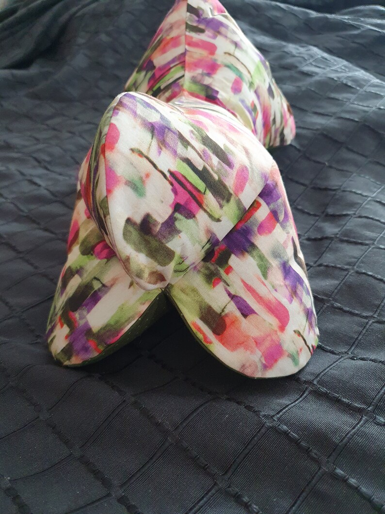 Op de afbeelding: Een kleurrijk kussen met een abstract patroon en een unieke, gebogen vorm. Het kussen is gemaakt van een zachte, comfortabele stof en heeft een witte achtergrond met roze, paarse, groene en zwarte accenten.