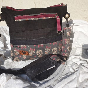 Könnte beinhalten: Schwarze Canvas-Umhängetasche mit einem pinkfarbenen und grauen Totenkopf-Druck. Die Tasche hat eine Reißverschlusstasche und einen langen verstellbaren Riemen.
