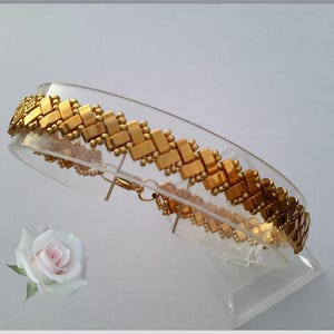 Peut inclure: Un bracelet de couleur dorée présentant un motif géométrique tissé de perles rectangulaires, agrémenté de petites perles rondes sur les bords. Le bracelet est présenté sur un support incurvé transparent, mettant en valeur son design.