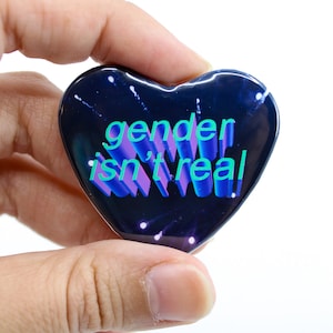 May include: A heart-shaped button with a black background and white stars. The text "gender isn't real" is written in a 3D style with blue, green, and purple letters.