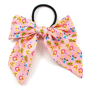 May include: Pink hair tie with a large bow. The bow is made of pink fabric with a repeating pattern of colorful hearts, swords, and crowns.