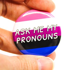 May include: A round button with the text "Ask Me My Pronouns" in white lettering. The button has a pink, white, purple, black, and blue striped design.