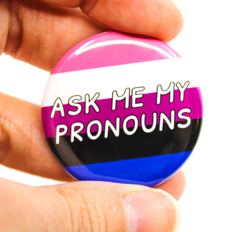 May include: A round button with the text "Ask Me My Pronouns" in white lettering. The button has a pink, white, purple, black, and blue striped design.